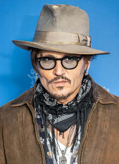 Johnny Depp wearing a beige fedora, round glasses, and a brown suede jacket against a blue background
